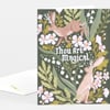 A folded greeting card with a dark green floral design featuring two rabbits and the centered white text "Thou Art Magical."