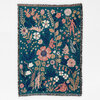 A rectangular woven blanket or throw with fringe edges and a teal floral pattern featuring stylized flowers, leaves, and several small human figures.