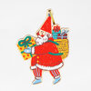A Santa Claus ornament cutout with a gold hanging ribbon, wearing a red suit and hat and carrying three colorful wrapped presents.