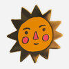A stylized sun enamel pin with a black spiky outline, an orange face, pink cheeks and nose, and simple black facial features.