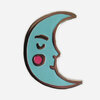 A crescent moon enamel pin with aqua blue fill, brown metal outline, a simple face, and a pink circular cheek detail.