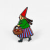 A colorful enamel pin of a little witch in a red pointed hat, green top, striped skirt, and teal shoes carrying a basket of apples.