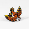 An enamel pin shaped like a small brown and yellow owl with spread wings and a white face.