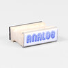 A small rubber stamp with a rectangular wooden block and a clear front face printed with the word "ANALOG" in blue stylized lettering.