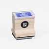 A small wooden rubber stamp with a clear top showing a blue label, black foam base, and a black circular side sticker with a white "P".