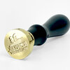 A brass wax seal stamp with a black wooden handle and a circular engraved head showing a snail design and mirrored lettering.
