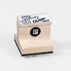 A wooden rubber stamp with a black foam base, a printed top label showing a cartoon figure and the words "DUMP DEJOY," and a black circle logo with a white "P" on the front.