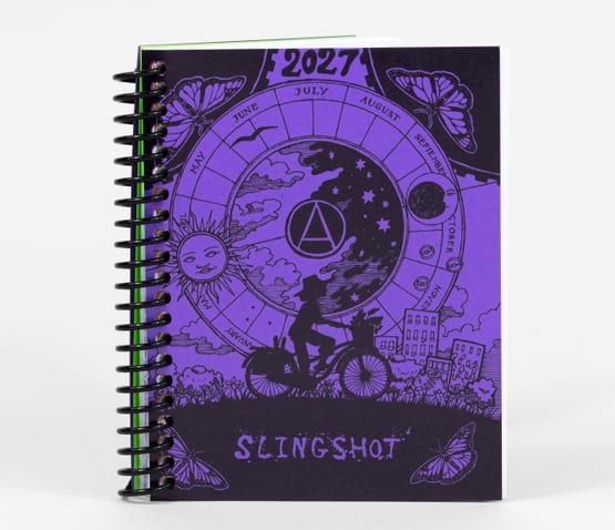 A mini spiral-bound planner with an orange and black illustrated cover showing "2027," a circular calendar design with month labels, butterflies, a sun face, a cyclist silhouette, and the word "SLINGSHOT."
