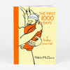 A small hardcover journal with a pale yellow cover, orange spine, and an illustration of an adult holding a swaddled baby, titled "THE FIRST 1000 DAYS" and "a baby journal" with the author name "Nikki McClure."