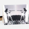 A folded greeting card featuring a black-and-white image of two women in matching white outfits aiming long-barreled guns at each other, with two dogs lying in the foreground.