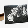A black-and-white greeting card shows a cat reaching toward a moon in a starry sky.