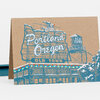 A folded greeting card with a kraft-brown front printed in blue and white with a stylized Portland, Oregon Old Town sign and building illustration, plus small multicolored dots along the roofline.