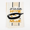 A cream-colored cloth with vertical black and mustard stripes and a skillet illustration, printed with the words "It Tastes Better HOME MADE".