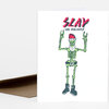 A folded greeting card with the text "SLAY THE HOLIDAYS" and a cartoon skeleton wearing a Santa hat, belt, and boots with raised arms.