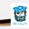 A folded greeting card with a pirate face illustration in blue and dark green and the text "Me Loves Ye" on the front.