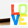 A folded greeting card or note card with the word "LOVE" in large hand-painted block letters in red, green, blue, and yellow on a white front.