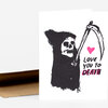 A white greeting card or folded card featuring a black grim reaper illustration holding a scythe, a pink heart, and the text "LOVE YOU TO DEATH" in black and pink.