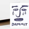 A folded white greeting card with a bold dark blue face illustration and the word "DAMMIT" printed at the bottom.