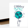 A folded greeting card with the hand-lettered text "CONGRATS YOU MAKE IT LOOK EASY" above a turquoise globe balanced on a raised finger illustration.