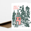 A folded holiday card with the text "HAVE A COZY HOLIDAY" and a hand-printed illustration of a small red house surrounded by tall evergreen trees.