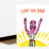 A folded greeting card with the text "LOVE YOU DAD" above a colorful illustration of a bearded shirtless man raising both hands over a yellow sun shape.