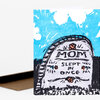 A folded greeting card with an illustrated tombstone on the front that reads “MOM SLEPT IN ONCE,” with small floral accents and a bright blue sky-like background.