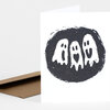 A folded white greeting card with a black circular print of three cartoon ghosts, one with a heart shape on its body, and a plain brown envelope behind it.