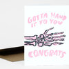 A folded card with pink lettering that reads "GOTTA HAND IT TO YOU" and "CONGRATS" above a black-and-pink skeleton hand illustration.