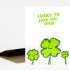 A folded greeting card with the handwritten text "LUCKY TO HAVE YOU DAD" and three smiling green clover illustrations on the front.
