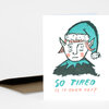 A folded greeting card with an illustrated tired elf wearing a teal hat and the text "SO TIRED IS IT OVER YET?" on the front, shown with a brown envelope.