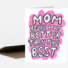 A folded greeting card with hand-drawn black-and-white bubble lettering over a bright pink shape that reads "MOM YOU'RE WAY BETTER THAN THE BEST."