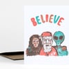 A folded greeting card with the word “BELIEVE” in multicolored letters above a stylized illustration of Santa between a Bigfoot-like figure and a green alien.