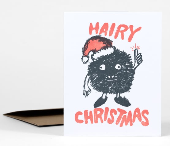 A folded holiday card shows a furry black cartoon character wearing a Santa hat and pointing upward, with the words "HAIRY" and "CHRISTMAS" in large coral letters.