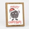 A kraft paper box set labeled "SIX LETTERPRESS PRINTED CARDS" and "w/ KRAFT ENVELOPES," with a front card showing the words "HAIRY CHRISTMAS" and a fuzzy black character wearing a Santa hat and holding up one finger.