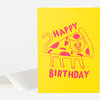 A folded yellow birthday card with a pink line drawing of a cat in a pizza slice and the words "HAPPY BIRTHDAY" on the front.